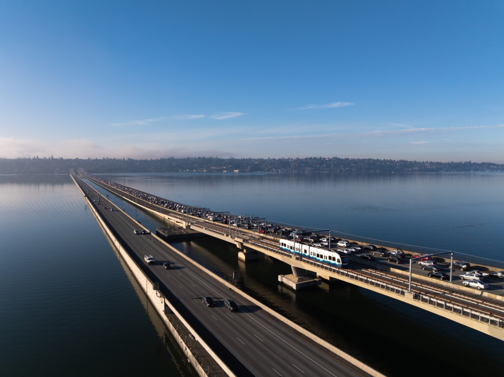Aerial view of a floating bridge spanning a large body of water, with light rail tracks and heavy vehicle traffic crossing under a clear blue sky.